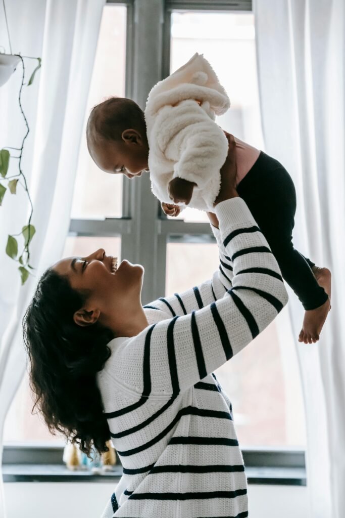 pexels-photo-6393374-6393374 Side view of cheerful African American mother and cute black baby in hands looking at each other while standing near window
