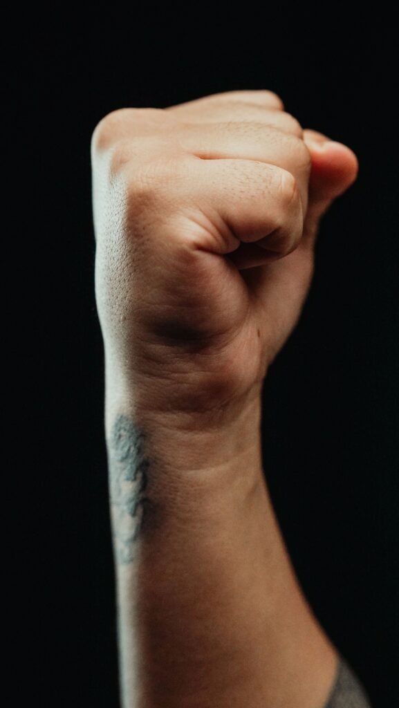 pexels-photo-7298919-7298919 Raised fist with tattooed arm on a dark background, symbolizing power or resistance.